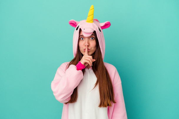 Giovane donna caucasica che indossa un pigiama unicorno isolato su sfondo blu mantenendo un segreto o chiedendo silenzio. - Foto, immagini