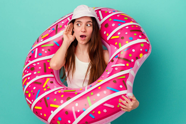 Young caucasian woman with an inflatable donut isolated on blue background trying to listening a gossip. - Photo, Image