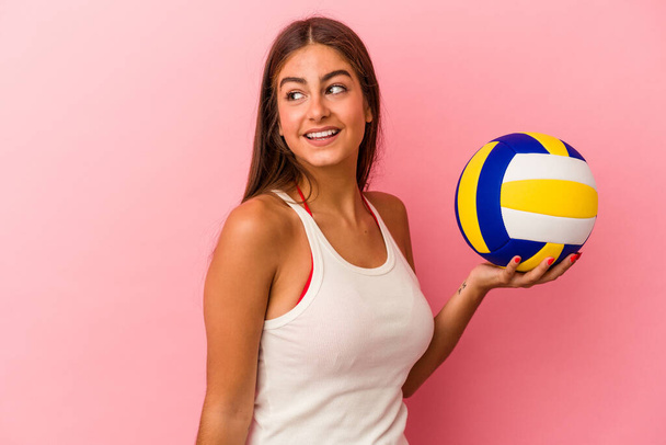 Fiatal fehér nő kezében röplabda elszigetelt rózsaszín háttér néz félre mosolygós, vidám és kellemes. - Fotó, kép