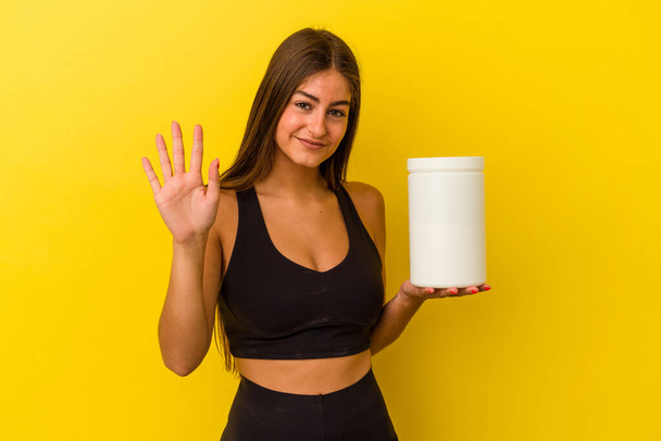 Jeune femme caucasienne tenant une bouteille de protéines isolée sur fond jaune souriant gai montrant numéro cinq avec les doigts. - Photo, image