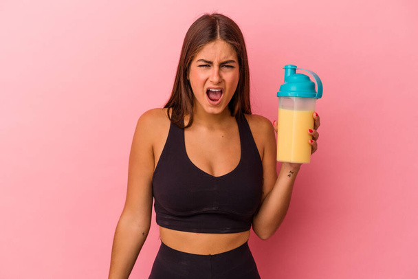 Young caucasian woman holding a protein shake isolated on yellow background screaming very angry and aggressive. - 写真・画像