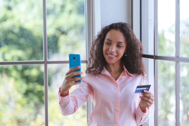 若いラテン語の女性は笑顔とオンラインショッピングのためのスマートフォンを使用しています。ビジネスの女性やフリーランスの仕事から家庭や電子決済のためのクレジットカードを保持します。オンラインストアでの幸せな買い物サービス. - 写真・画像