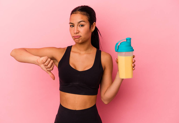 Young Venezuelan woman drinking a protein shake isolated on pink background showing a dislike gesture, thumbs down. Disagreement concept. - Photo, Image