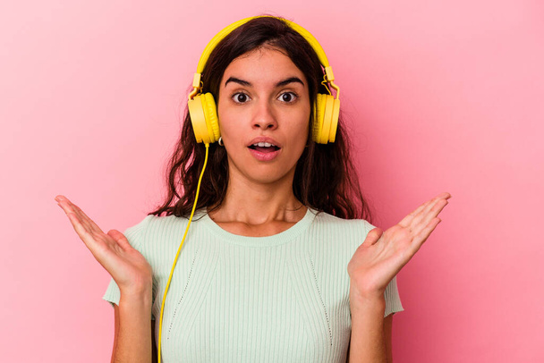 Junge kaukasische Frau hört Musik isoliert auf rosa Hintergrund überrascht und schockiert. - Foto, Bild