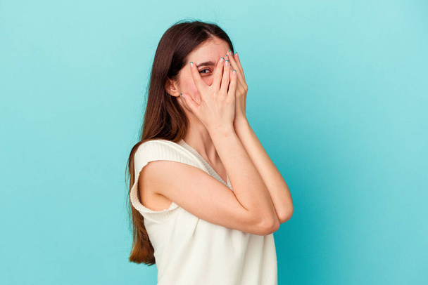 Jeune femme caucasienne isolée sur fond bleu cligner des yeux à la caméra par les doigts, visage couverture embarrassé. - Photo, image