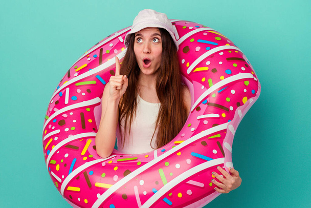 Mulher branca jovem com um donut inflável isolado no fundo azul apontando de cabeça com a boca aberta. - Foto, Imagem