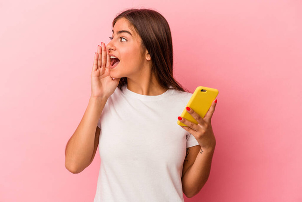 Nuori valkoihoinen nainen pitelee matkapuhelinta, joka on eristetty vaaleanpunaiselle taustalle huutaen ja pitäen kämmentä lähellä avattua suuta. - Valokuva, kuva
