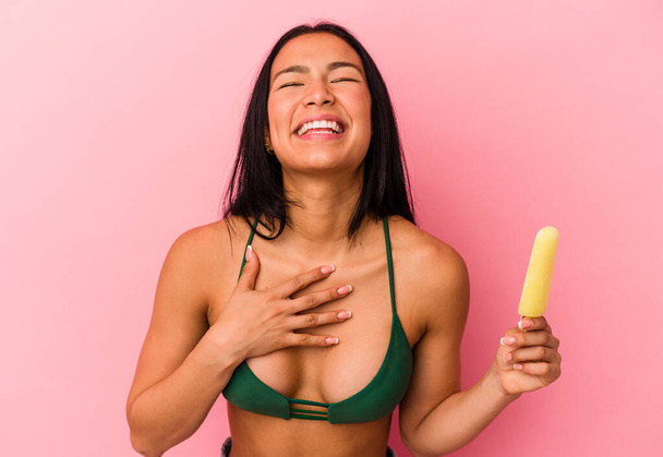 Молодая венесуэльская женщина, держащая мороженое на розовом фоне, громко смеется, держа руку на груди.. - Фото, изображение