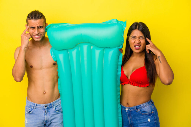 Joven pareja latina sosteniendo colchón de aire aislado sobre fondo amarillo mostrando un gesto de decepción con dedo índice. - Foto, imagen