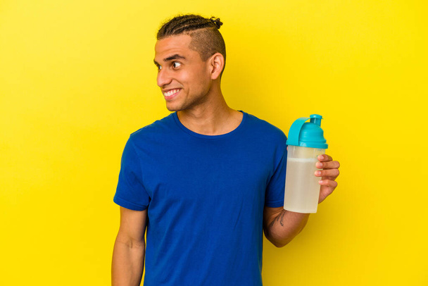 Joven venezolano bebiendo un batido de proteínas aislado sobre fondo amarillo mira a un lado sonriente, alegre y agradable. - Foto, Imagen