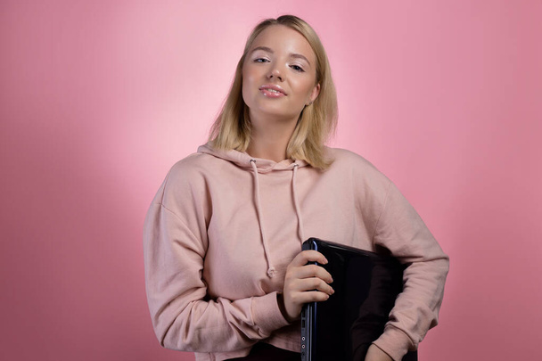young woman with a laptop, a student or a freelancer. A charming blonde in a pink hoodie - Fotó, kép