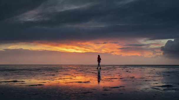 Kobieta sylwetka spaceru wzdłuż Morza Wadden w Buesum podczas zachodu słońca, odbicie w mokrym piasku, Niemcy - Zdjęcie, obraz