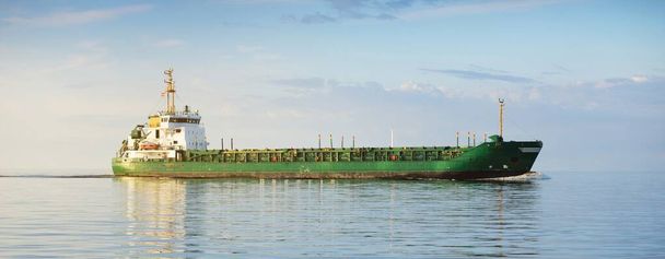 Великий зелений вантажний корабель, що плаває у відкритому морі на заході сонця. Барвисті сяючі хмари, тепле сонячне світло. Зображення концепції. Вантажні перевезення, бізнес, економіка, промисловість, глобальні комунікації, логістика
 - Фото, зображення