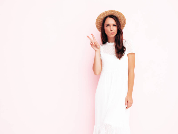 Young beautiful smiling female in trendy summer  clothes. Sexy carefree woman posing near pink wall in studio. Positive brunette model having fun. Cheerful and happy. In hat. Shows peace sign - Foto, imagen