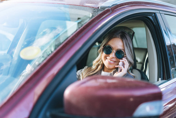 Giovane donna elegante con lunghi capelli biondi e occhiali da sole alla guida di una macchina e utilizzando il telefono cellulare - Foto, immagini