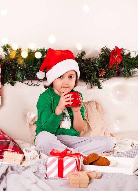 Little girl in funny Santa cap and Christmas pajamas sitting on a bed eating cookies and drinking cocoa - Fotografie, Obrázek