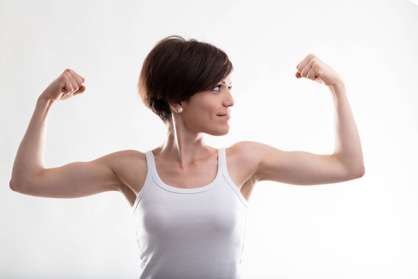 Pretty young woman wearing a sleeveless top while flexing her arms for showing her biceps muscles as a gesture of power and confidence against white background for copy space - Fotografie, Obrázek