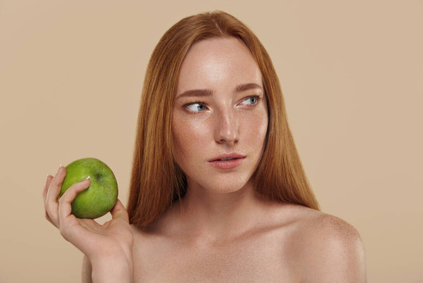 Redhead caucasian girl with naked shoulders holding apple. Cropped partial view of young beautiful red-haired woman with freckles. Natural female beauty. Isolated on beige background in studio. - Foto, imagen
