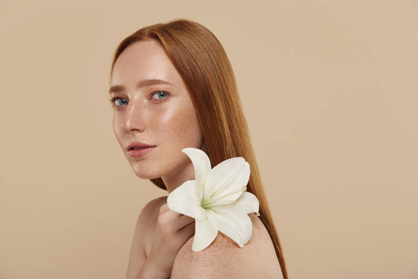 Redhead caucasian girl with naked shoulders holds lily flower. Cropped view of young beautiful red haired woman with freckles. Concept of natural female beauty. Isolated on beige background in studio - Photo, Image