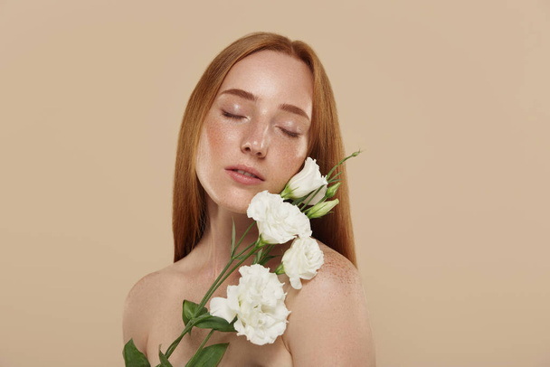 Caucasian girl with naked shoulders holds eustoma flower. Cropped partial view of young beautiful red haired woman with freckles. Concept of natural female beauty. Isolated on beige background - Photo, image