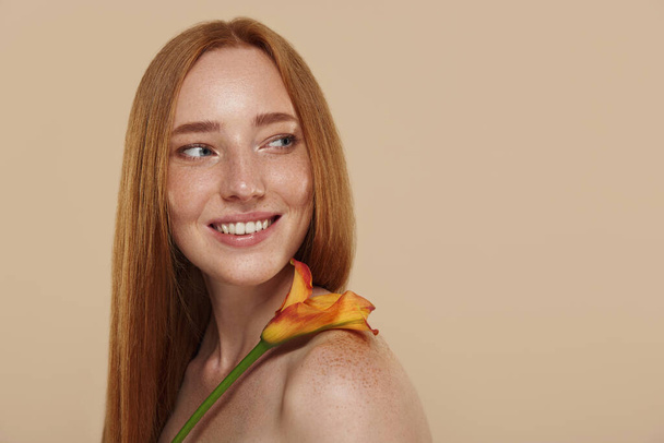 Redhead caucasian girl with naked shoulders holds calla flower. Cropped partial view of young beautiful red haired woman with freckles. Concept of natural female beauty. Isolated on beige background  - Φωτογραφία, εικόνα