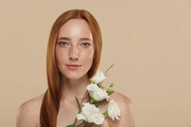 Caucasian girl with naked shoulders holds eustoma flower. Cropped partial view of young beautiful red haired woman with freckles. Concept of natural female beauty. Isolated on beige background - Photo, Image