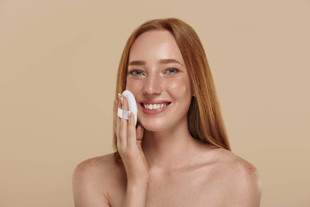 Redhead smiling caucasian girl with naked shoulders and powder puff. Cropped partial view of young beautiful red-haired woman with freckles. Natural beauty. Isolated on beige background in studio - Photo, Image