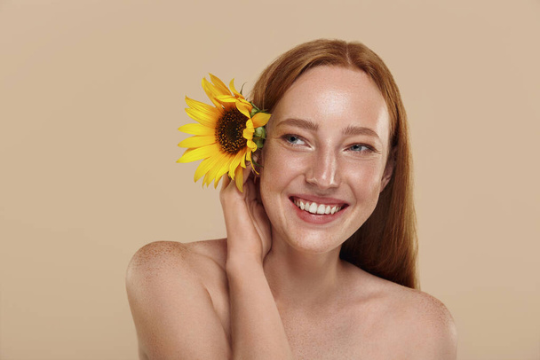 Redhead caucasian girl with naked shoulders holds sunflower. Cropped partial view of young beautiful red-haired woman with freckles. Concept of natural female beauty. Isolated on beige background. - Фото, изображение