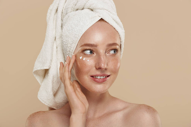 Caucasian girl with naked shoulders, undereye patches and towel on head. Cropped partial view of young beautiful woman with freckles. Natural female beauty. Isolated on beige background. - Zdjęcie, obraz