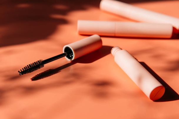 pink clean makeup brush for mascara lies next to an open tube, closed tubes of cosmetics, lip gloss, liquid lipstick, pink eyeliner on a peach background with shadows. Copy space - Photo, Image