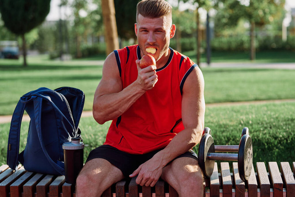 man in the park sits on a bench and eats an apple summer - Фото, зображення