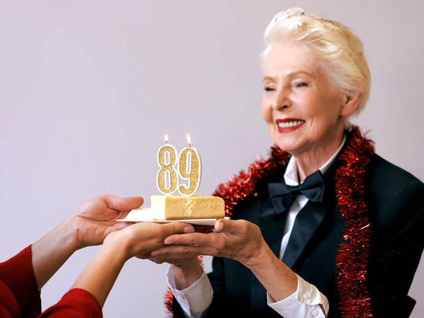 ケーキと一緒に誕生日を祝う黒いスーツの幸せな陽気なスタイリッシュな89歳の女性。ライフスタイル、ポジティブなファッション、スタイルコンセプト - 写真・画像
