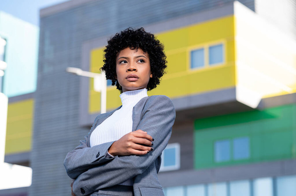 A cool young African American lady with curly hair dressed professionally posing near buildings - Фото, зображення