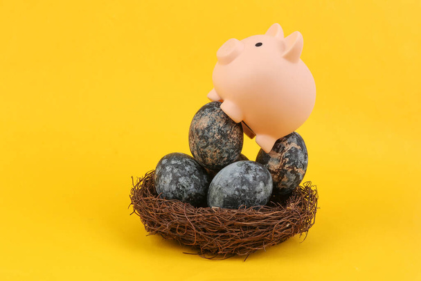 Prasátko banka sedí na šedém mramoru malované velikonoční vejce v hnízdě na žlutém pozadí - Fotografie, Obrázek