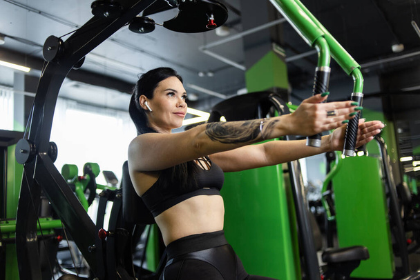 Young fitness woman exercising pectoral muscles doing hand reduction in exercise machine in gym - Photo, Image