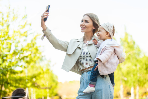 Милая мама с маленькой дочкой на руках делает селфи на смартфоне в парке - Фото, изображение