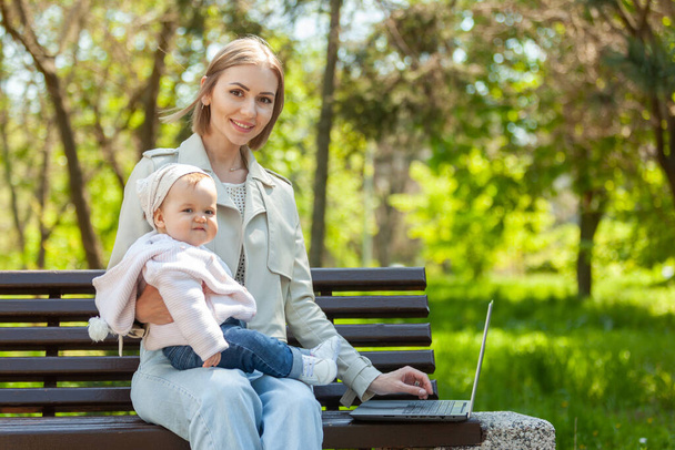 Tyylikäs liikeäiti käyttää kannettavaa tietokonetta vauva sylissään istuessaan penkillä puistossa. Etätyö, äitiys, äitiyslomalla oleva työ - Valokuva, kuva