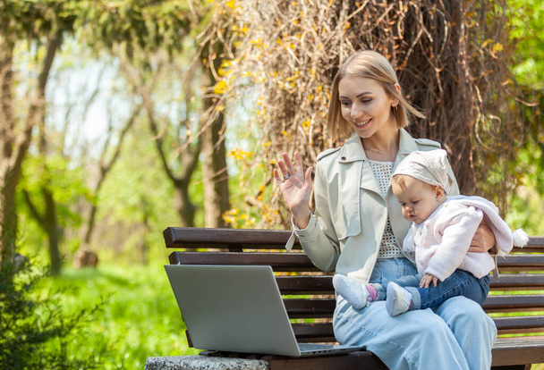 Tyylikäs liikeäiti tekee videopuhelun kannettavalla tietokoneella ja hänen tyttärensä sylissään istuessaan penkillä puistossa. Etätyö, äitiys, äitiyslomalla oleva työ - Valokuva, kuva