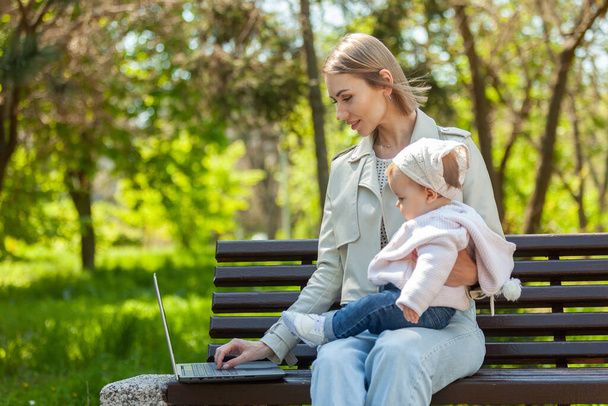 Stylish business mom uses a laptop with a baby in her arms while sitting on a bench in the park. Distance work, motherhood, work on maternity leave - 写真・画像