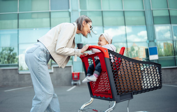 A caring mother with her little daughter is shopping. Lovely lady with baby and shopping trolley in parking - Foto, immagini