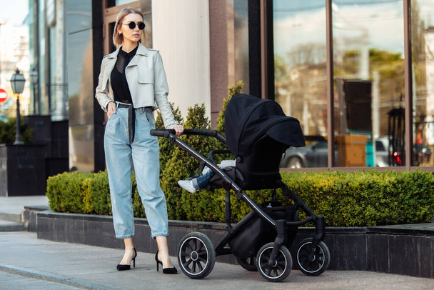 Стильная любящая мама гуляет с коляской в городе. Материнство, образ жизни - Фото, изображение