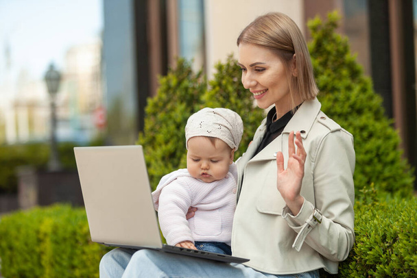 Biznesowa matka ze swoją córeczką wykonuje wideo rozmowy na laptopie w mieście. Praca nad urlopem macierzyńskim, macierzyństwem, stylem życia. Wolny strzelec, praca zdalna - Zdjęcie, obraz