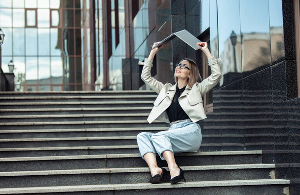 Business lady shakes laptop over her head looking for information while sitting on stairs outdoors. Education, work, freelance - Photo, Image