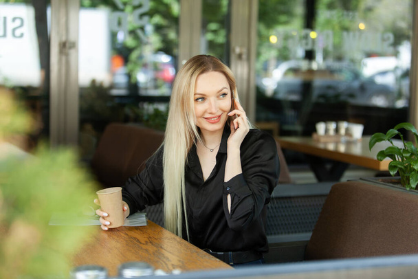 Young attractive blonde woman talking on the phone and drinking coffee while sitting in an outdoor cafe - Φωτογραφία, εικόνα