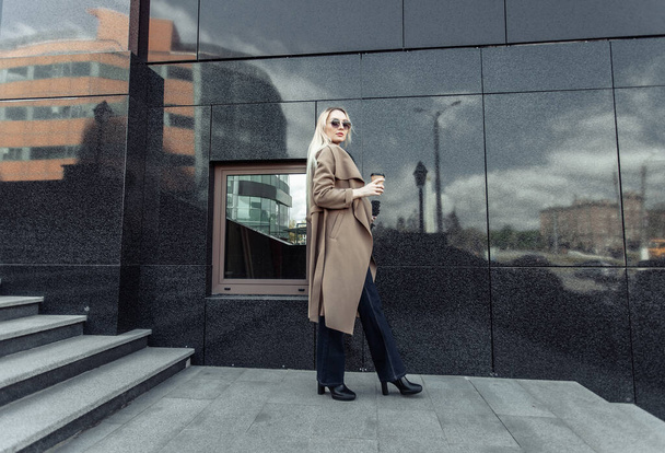 Blonde woman in trench coat holds cup of coffee in urban location - Photo, Image