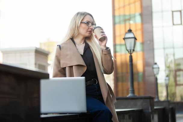 Bizneswoman używa laptopa i trzyma filiżankę kawy w miejskiej lokalizacji. Odległe, praca online - Zdjęcie, obraz