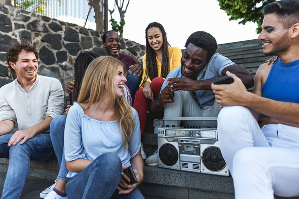 Молодые многорасовые друзья с удовольствием слушают музыку с винтажным магнитофоном, сидя на городской лестнице - концепция молодежного стиля жизни тысячелетия - Фото, изображение