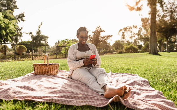 Mujer embarazada africana usando teléfono inteligente móvil mientras hace un picnic en el parque - Concepto de estilo de vida de maternidad - Foto, imagen