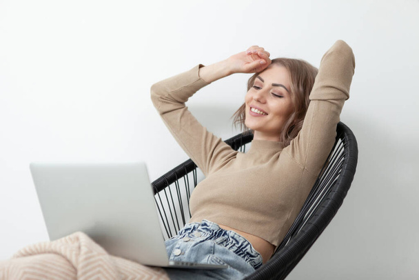Donna sorridente siede su una poltrona con un computer portatile coperto da una coperta sullo sfondo di una parete bianca. Tempo libero, stile di vita concetto - Foto, immagini