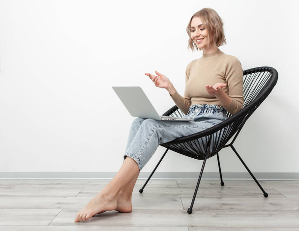 Cute blonde woman has video call or conference on laptop while sitting in chair against the backdrop of a white wall at home. Freelance, remote work concept - Photo, Image